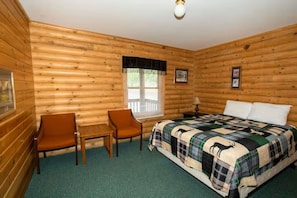 A log cabin room with a bed, two chairs, a small table, a window with blinds, and a ceiling light.