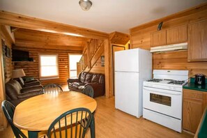 LâintĂ©rieur dâune cabane en rondins comprend un coin repas, une cuisine Ă©quipĂ©e dâun poĂȘle et dâun rĂ©frigĂ©rateur, ainsi quâun escalier.