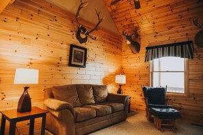 A log cabin interior with a brown leather sofa, two lamps, a framed picture, and a deer head mounted on the wall.
