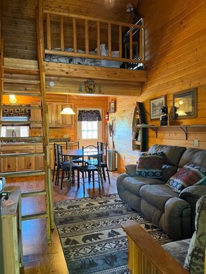 Ein Holzblockhaus-Interieur mit Essbereich, einer Couch, einer Treppe und einem Schlafzimmer im Obergeschoss.