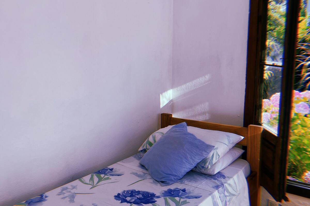 Quarto pequeno com uma cama de solteiro, cabeceira de madeira, edredom floral e janela com vista para o verde.