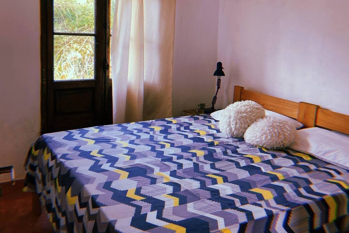 Quarto com cama, janela com cortinas e mesa de cabeceira com abajur.
