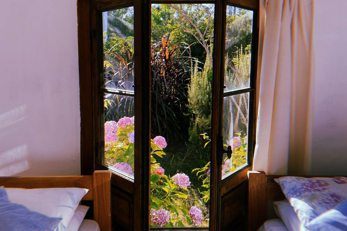 Quarto com duas camas, janela com cortinas e vista para um jardim com flores cor-de-rosa.