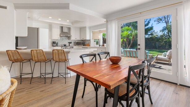 Une cuisine moderne avec des meubles blancs, une table à manger en bois et une vue sur un jardin, visible à travers des portes vitrées.