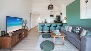 A modern living room with a TV, sofa, coffee table, and kitchen in the background. - 2CH Orient-Bay beach walk (Orient-Baie)