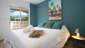 A bedroom with a bed, a painting of a beach scene, and a view of a balcony with a white fence. - 2CH Orient-Bay beach walk (Orient-Baie)