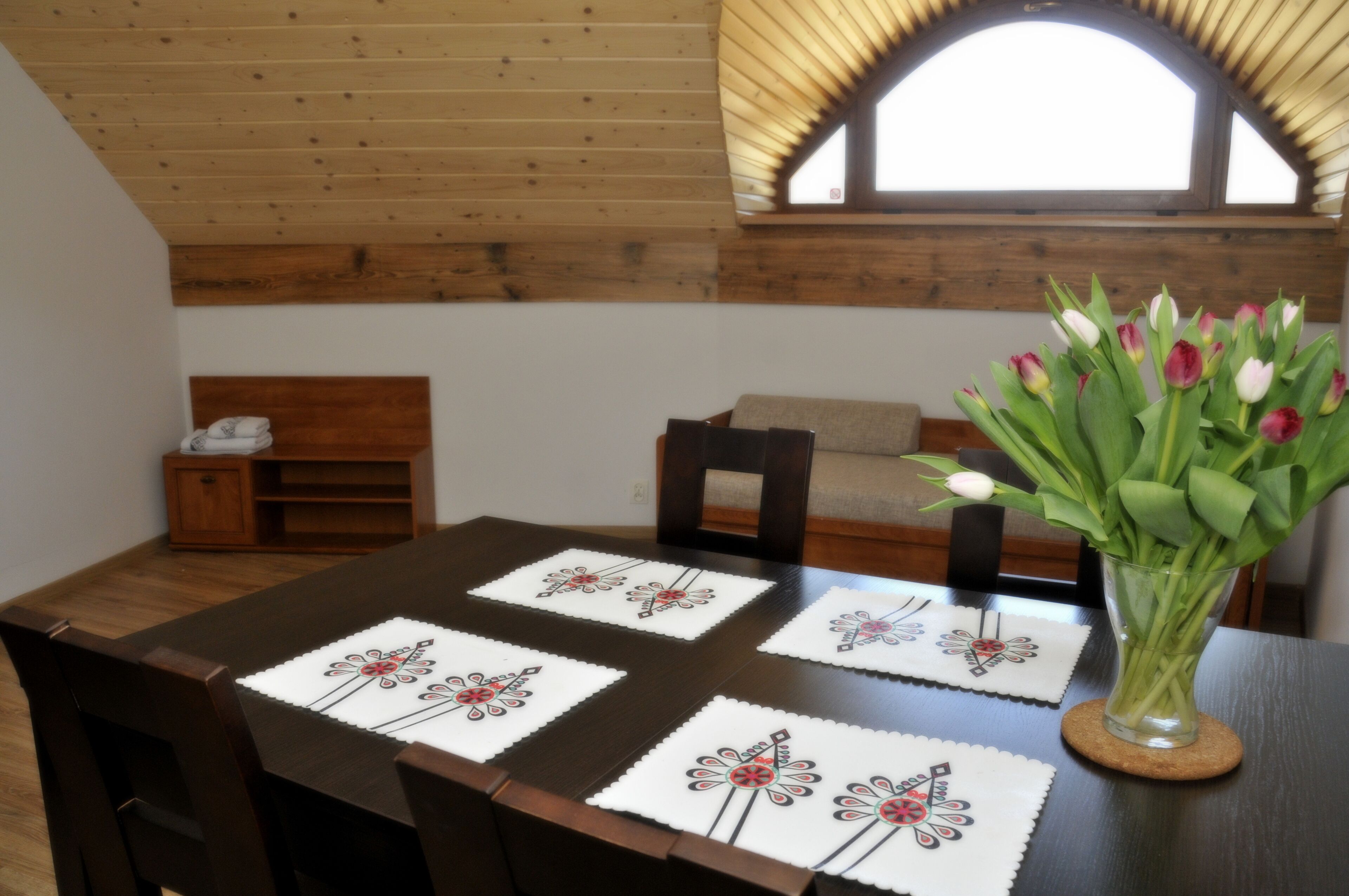 Une table à manger agrémentée de sets de table à motifs floraux, un vase de tulipes et un plafond en bois.