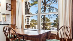 A dining area with a wooden table and chairs, large windows with curtains, and a view of trees and a building outside. - Bayview Beach Cottage For 4! (Saco)