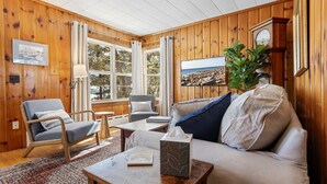 A cozy room with wooden walls, a sofa, a chair, a table, and a window with curtains. - Bayview Beach Cottage For 4! (Saco)