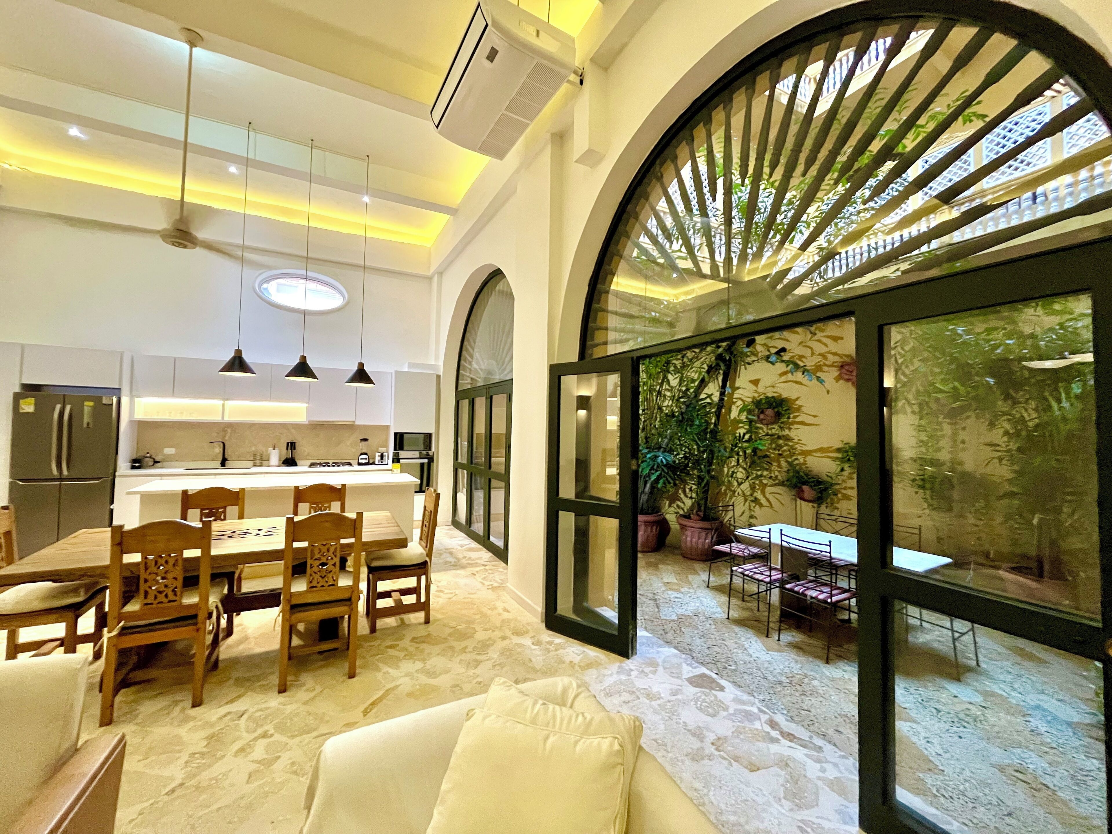 Cocina moderna con una isla central, comedor con sillas y un gran ventanal que da a un espacio exterior con plantas.