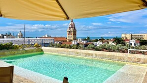 A swimming pool with a tiled edge, a wooden deck, and a view of a cityscape including a prominent building with a dome and bell tower. - by Calamari Homes - El Porvenir (Cartagena)