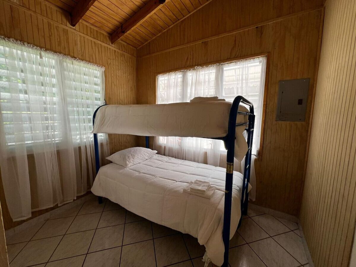 A bunk bed in a wooden room with tiled flooring and two windows with sheer curtains.
