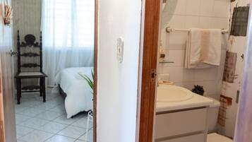 Une chambre à coucher avec un lit, une table de chevet et une chaise. La pièce est dotée d’une porte blanche et d’une fenêtre avec des rideaux.