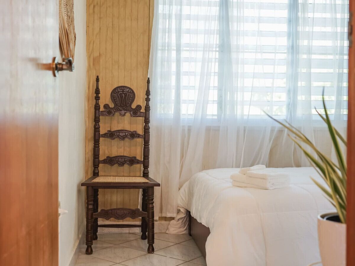 Una silla de madera con intrincados grabados, una cama con sábanas blancas y una planta en maceta en una esquina.