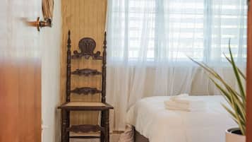 Una silla de madera con intrincados grabados, una cama con sábanas blancas y una planta en maceta en una esquina.