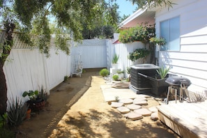 Un jardin avec une terrasse en bois, des plantes en pot, un barbecue et une clôture blanche.
