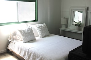 Un lit bien fait, avec une literie et des oreillers blancs, une lampe de chevet, un miroir au-dessus du lit et une fenêtre avec des stores.