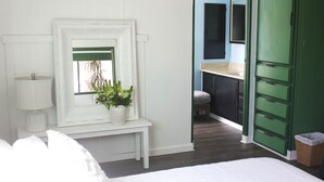 A bedroom with a bed, a bedside table with a lamp, a mirror, a potted plant, and a view into a bathroom with a sink and mirror.