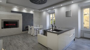 A modern kitchen with a built-in island, a dining area with white chairs, a flat-screen TV mounted on the wall, and a window providing natural light.