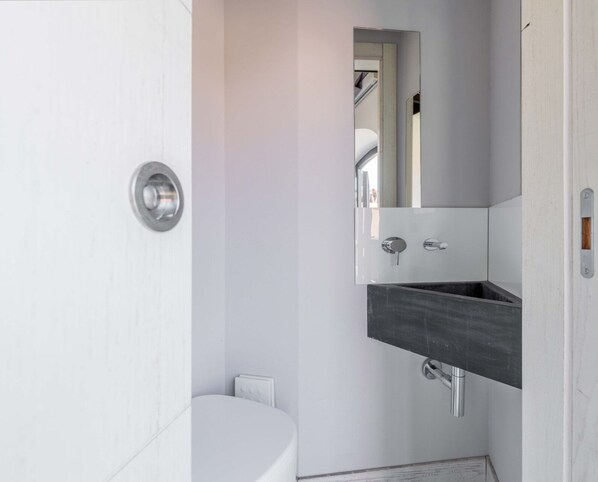 A compact bathroom with a white toilet, a wall-mounted sink with a dark countertop, and a mirror reflecting part of the room. - Mr. Brown - Attic La Grande Bellezza (Roma)