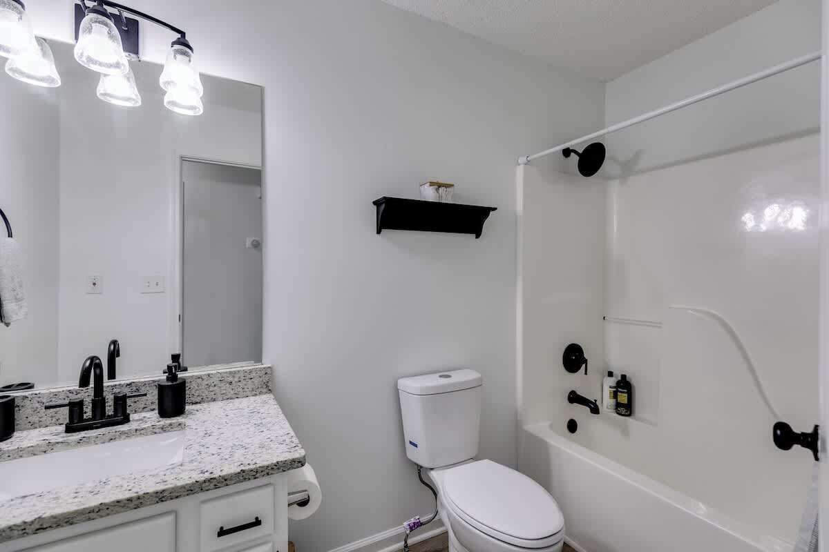 A bathroom with a white sink, black faucet, and a mirror with lights. There is a toilet and a shower area with a white tub.
