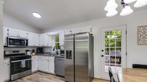 A modern kitchen with white cabinets, stainless steel appliances, and a dining area with a wooden table. - Pet-Friendly 3BR | Fenced Yard + Fire Pit (Villa Rica)