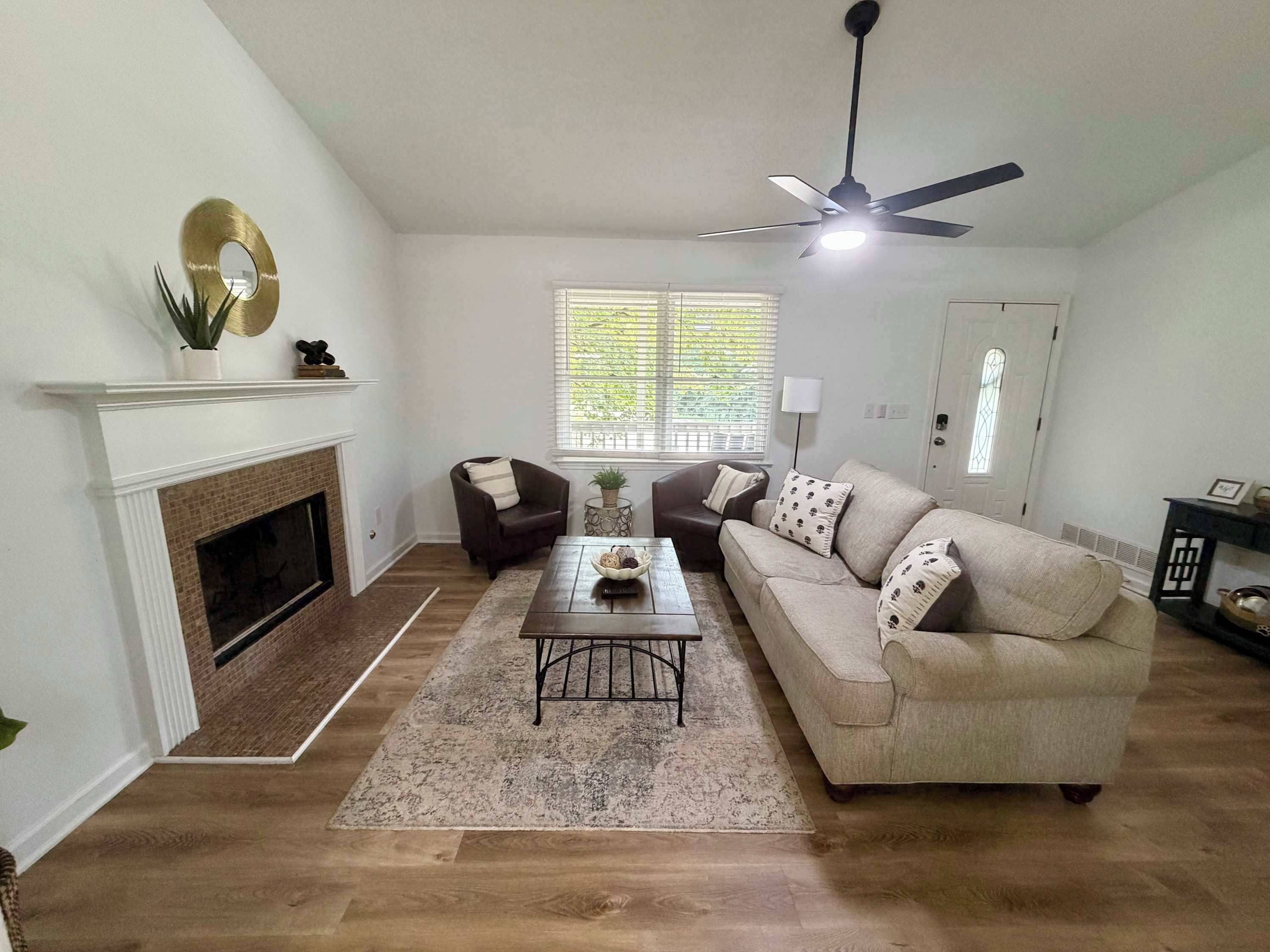 A living room with a fireplace, a ceiling fan, and a large window with blinds.
