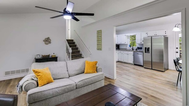 A modern living room with a grey sofa, wooden coffee table, and a ceiling fan. A staircase leads to a kitchen with white cabinets and a stainless steel refrigerator. - Pet-Friendly 3BR | Fenced Yard + Fire Pit (Villa Rica)