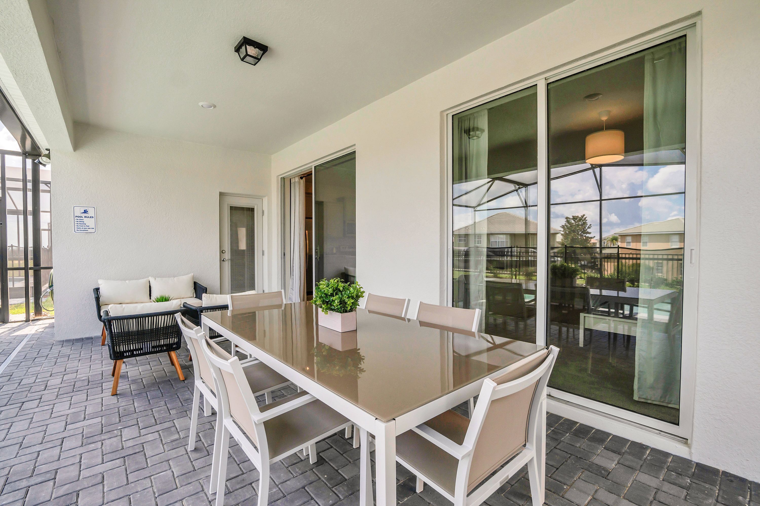 A modern outdoor dining area with a glass table and white chairs, a cushioned sofa, and a potted plant.
