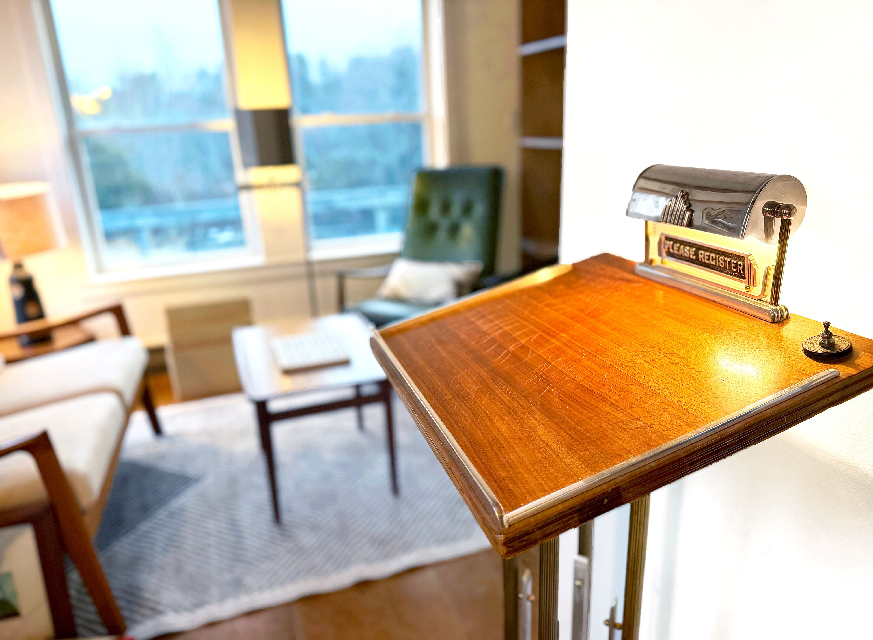 A wooden table with a registration sign, a laptop on a small table, a green chair, and a view of the city through large windows.