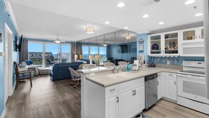 A modern kitchen with white cabinets, a dishwasher, and a large island with a sink. There is a dining area with a blue sofa and wicker chairs.