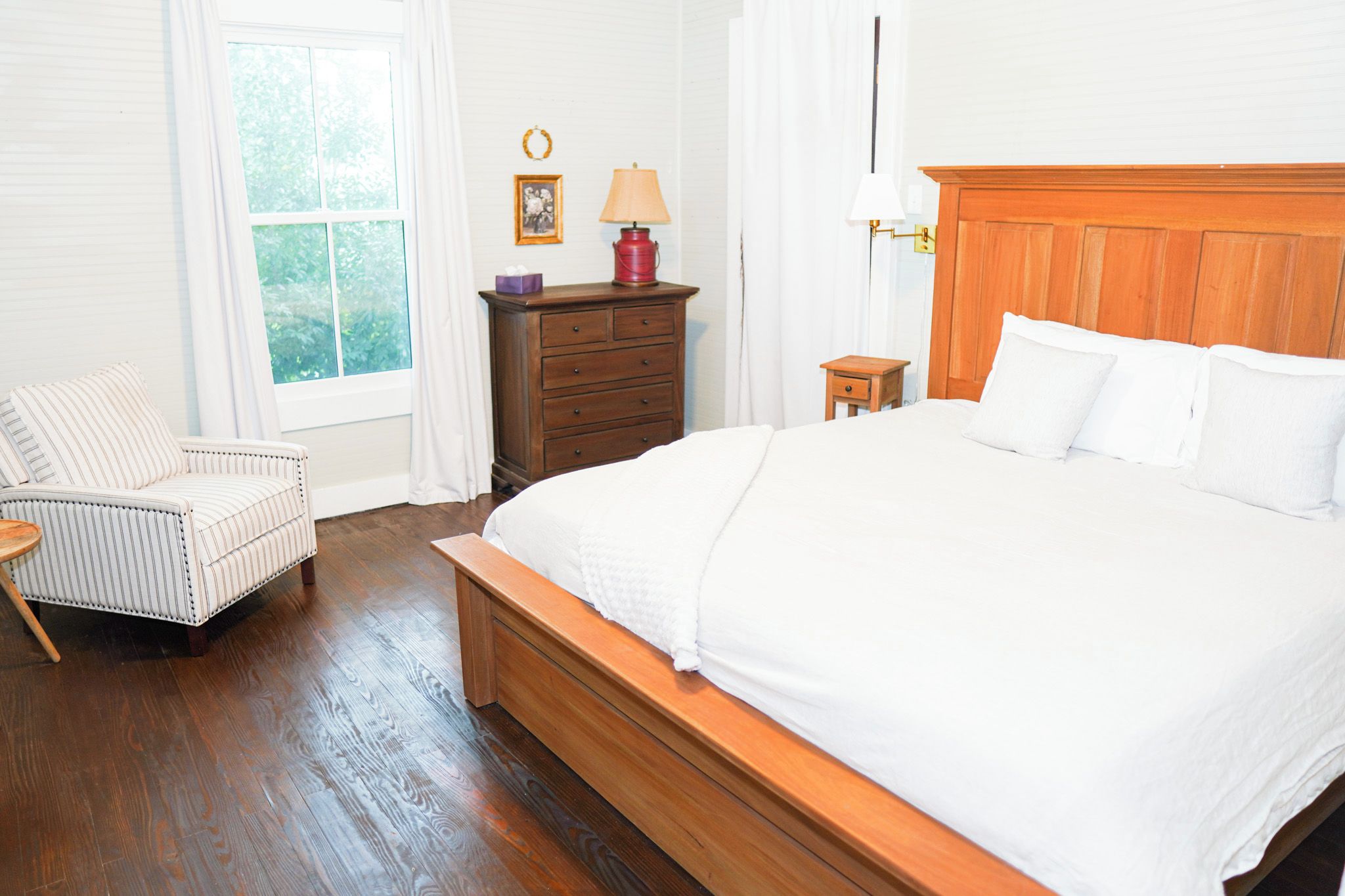 A bedroom with a large bed, a wooden headboard, a nightstand, a dresser, a chair, and a window with curtains.
