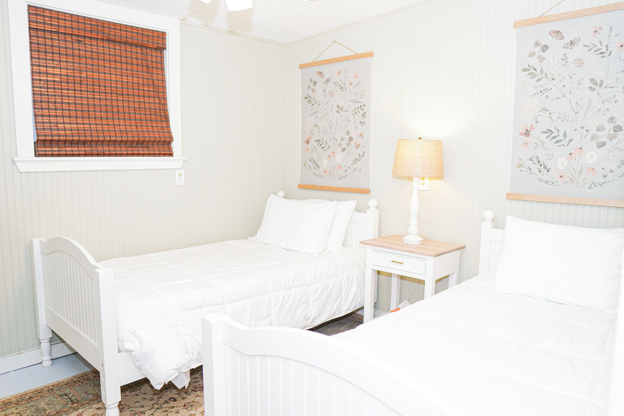 Una habitación con dos camas, una mesita de noche, una lámpara y decoración floral en la pared.