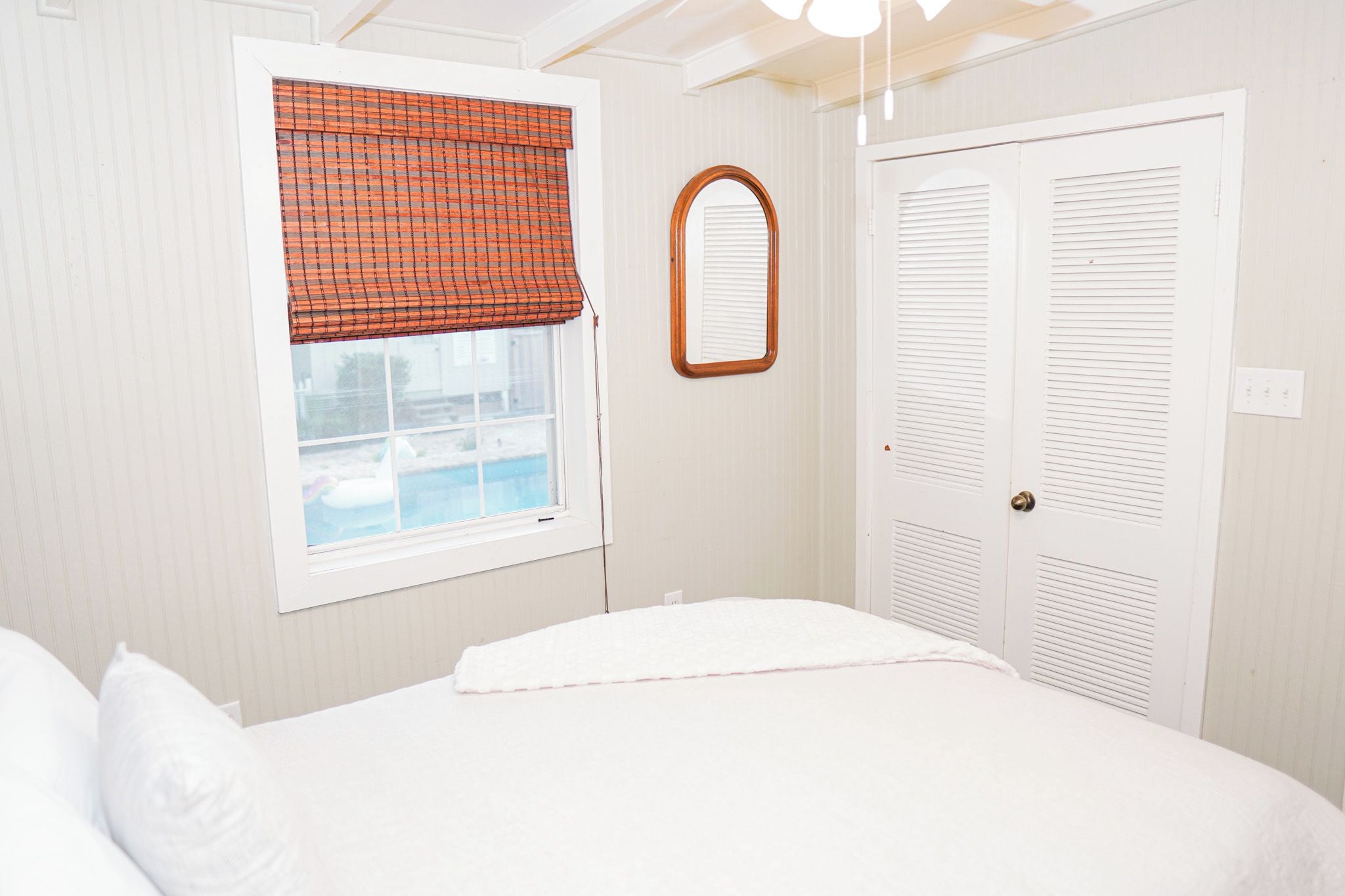 A bedroom with a bed, a window with a checkered blind, a mirror, and a set of double doors.