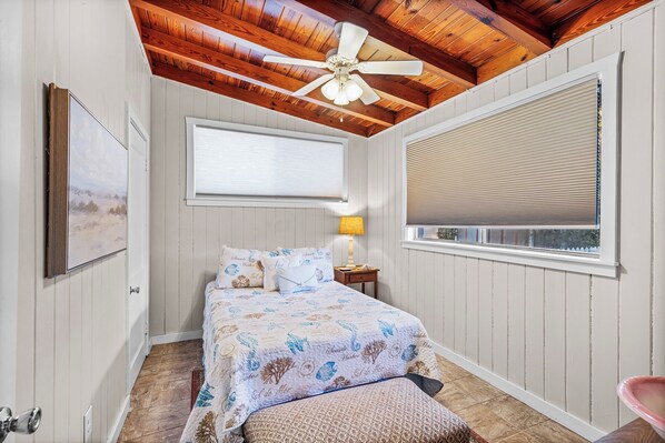 A bedroom with a bed, a ceiling fan, a window with blinds, a bedside lamp, and a framed picture on the wall.