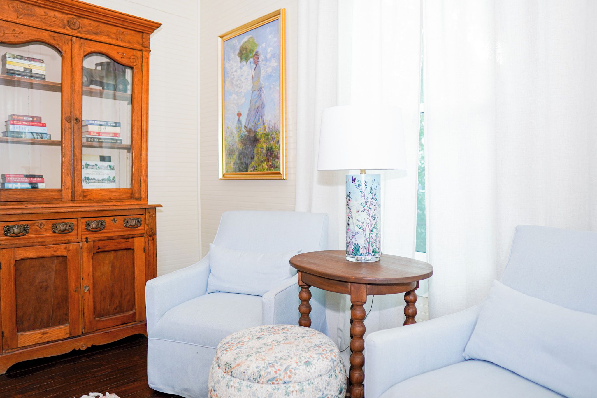 A room with a wooden cabinet, two white armchairs, a round wooden table, a floral ottoman, a painting, and a lamp.