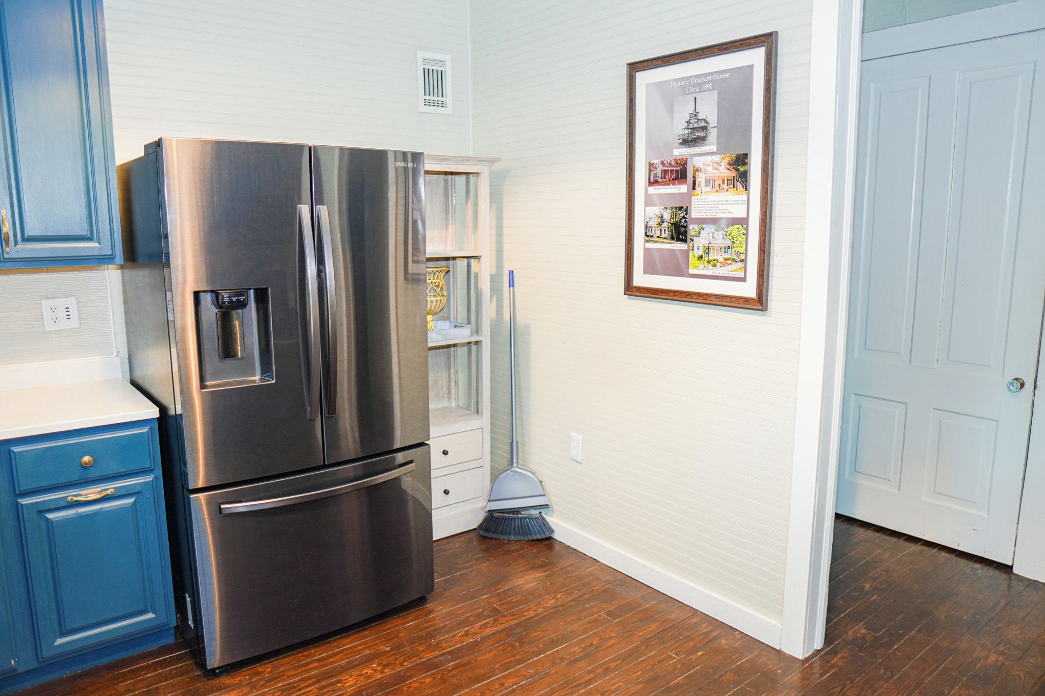 A kitchen with a stainless steel refrigerator, blue cabinets, a broom, and a framed picture on the wall.
