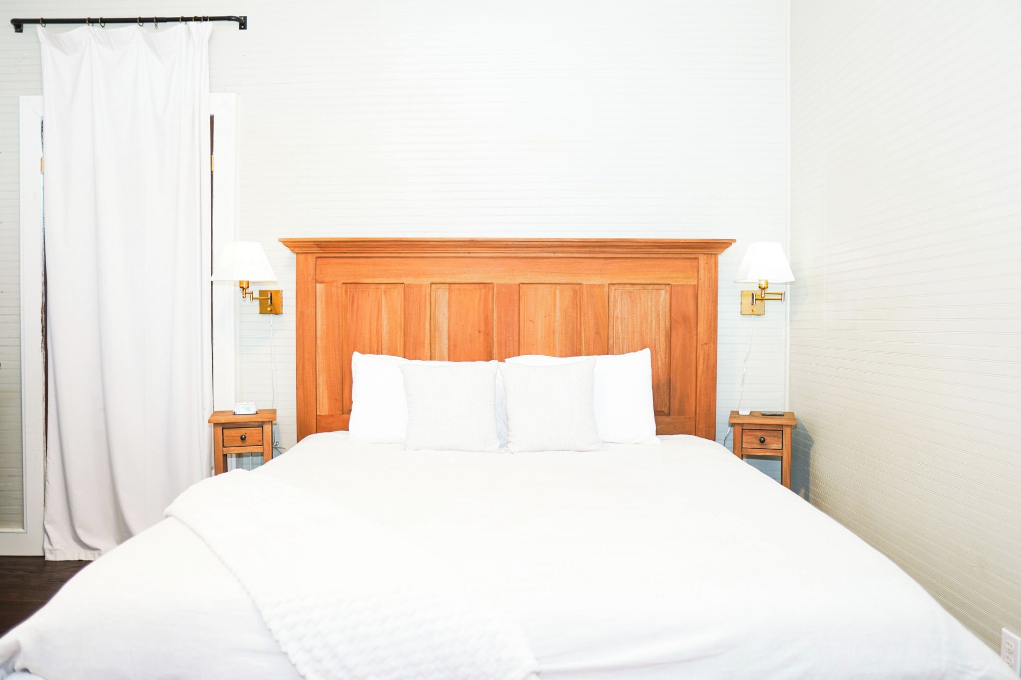 Une chambre d’hôtel avec une tête de lit en bois, du linge de lit blanc, deux tables de chevet avec des lampes et un rideau blanc.
