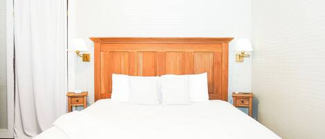 Une chambre d’hôtel avec une tête de lit en bois, du linge de lit blanc, deux tables de chevet avec des lampes et un rideau blanc.