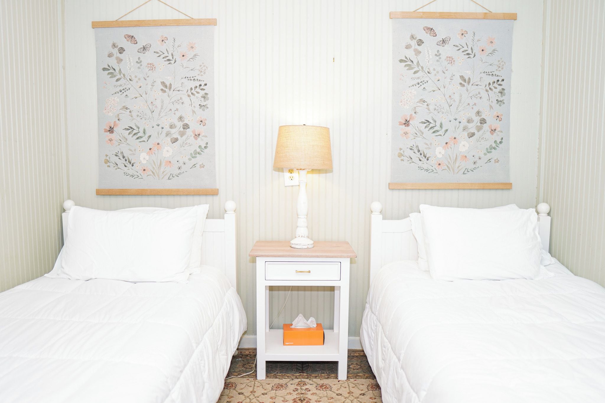 Ein Hotelzimmer mit zwei Einzelbetten, einem Nachttisch mit Lampe und zwei Blumenbildern an der Wand.
