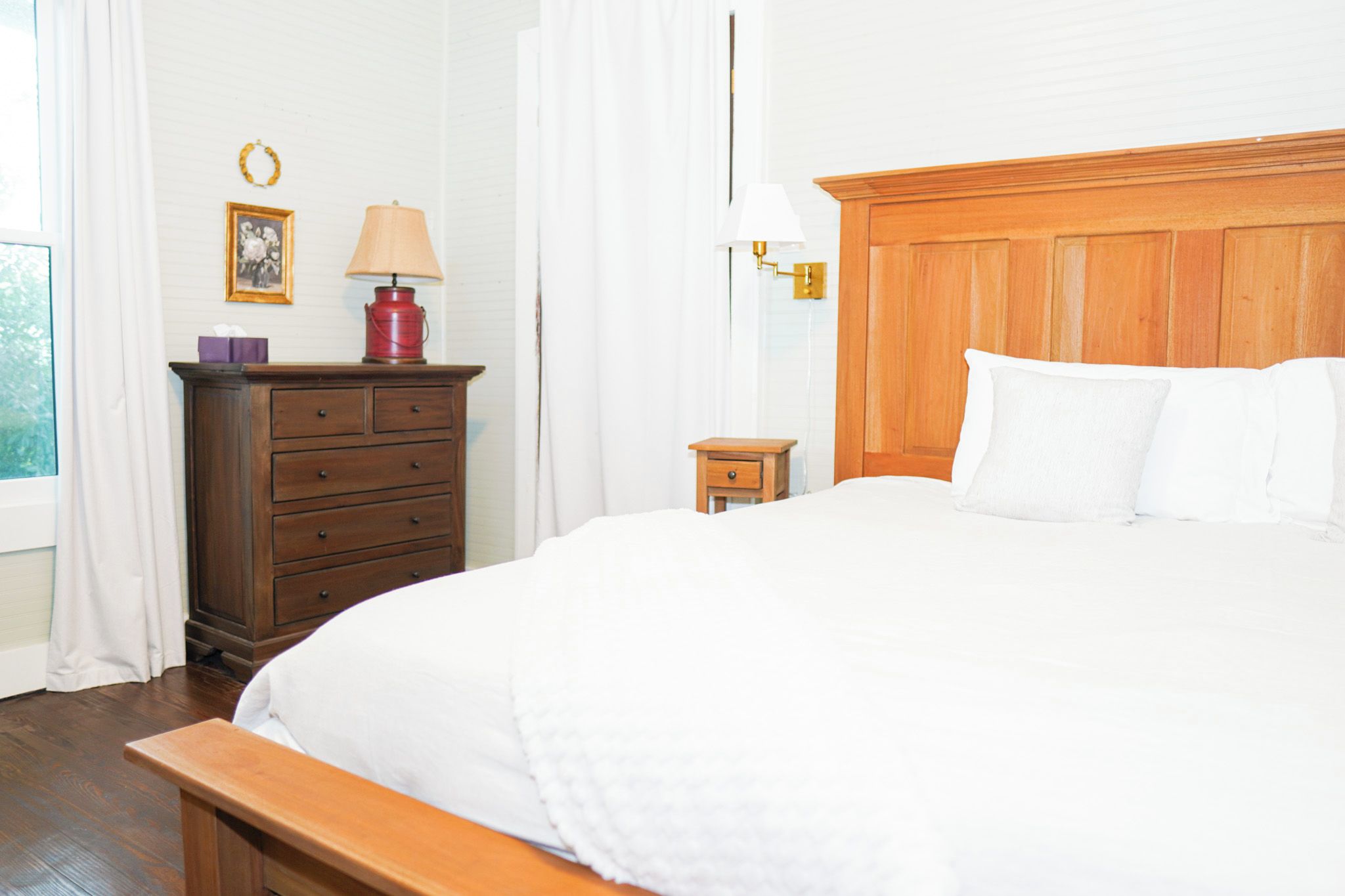 A neatly arranged bedroom with a wooden headboard, a bedside table with a lamp, a dresser, and a window with curtains.