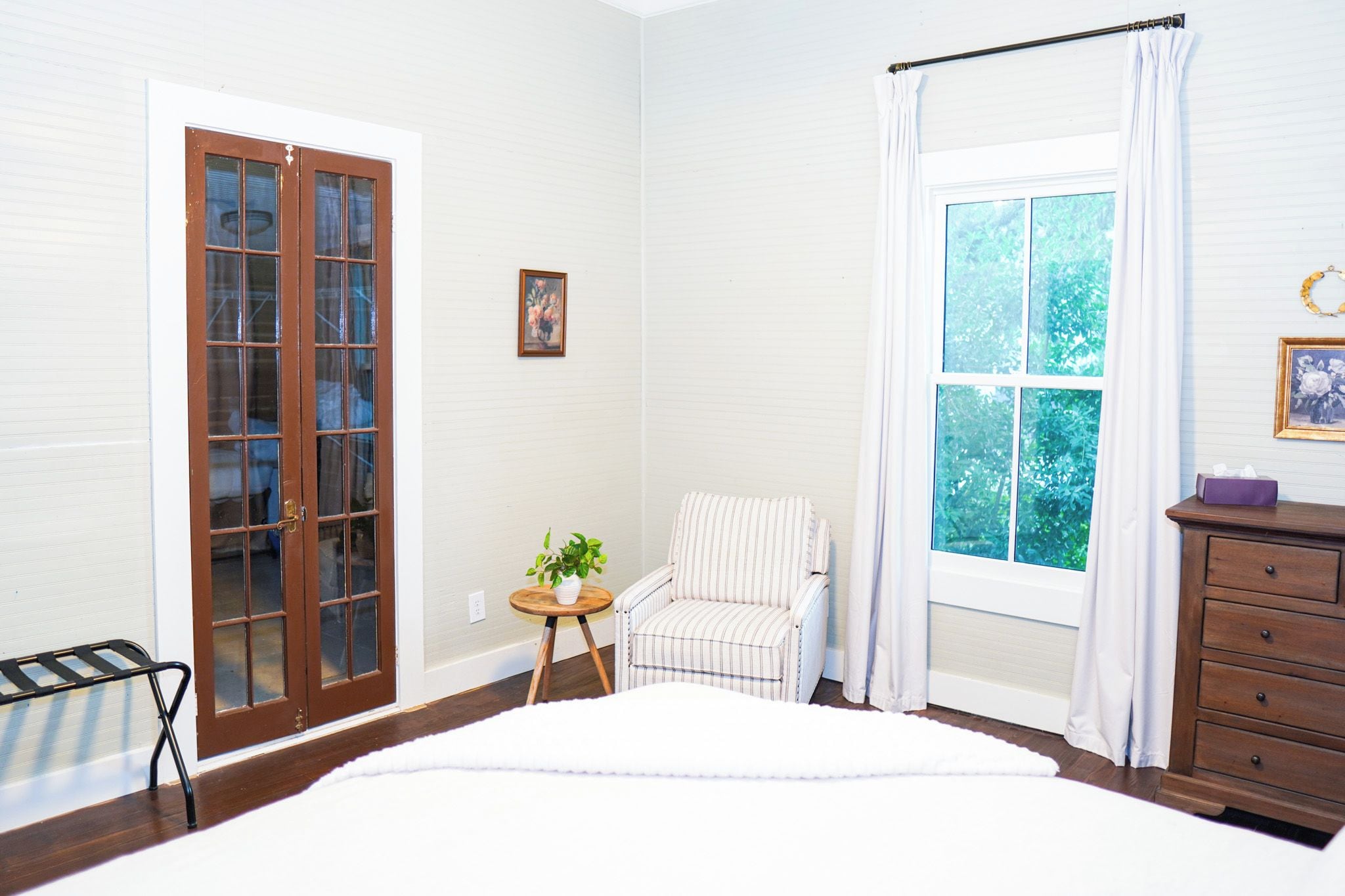 A bedroom with a large bed, a window with curtains, a chair, a small table with a plant, and a dresser.