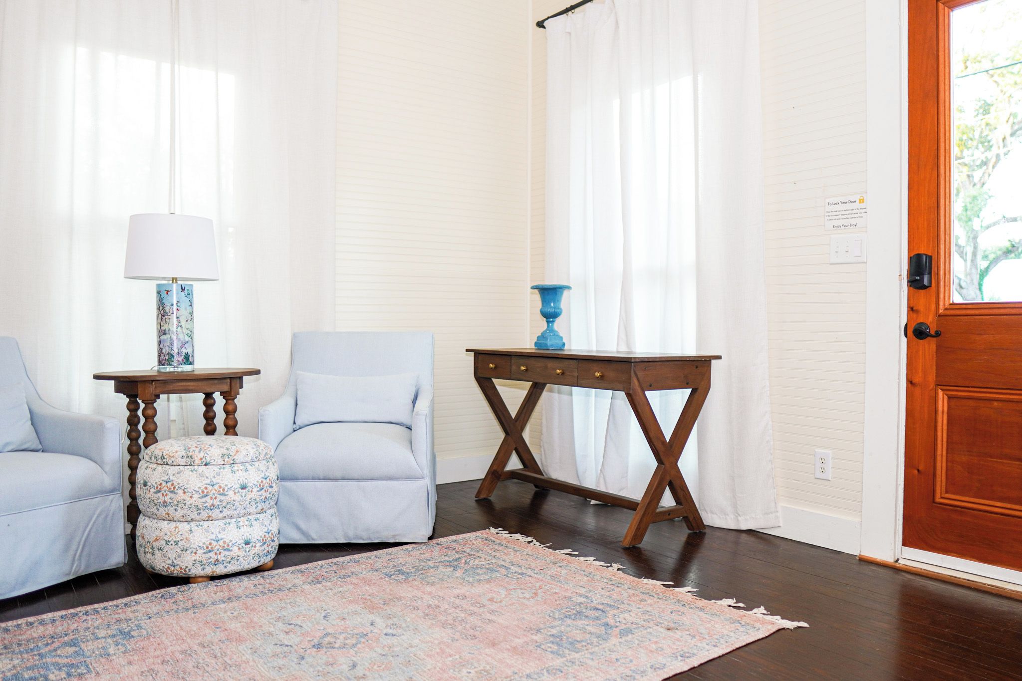 A room with a wooden door, two armchairs, a small table with a lamp, and a patterned rug.