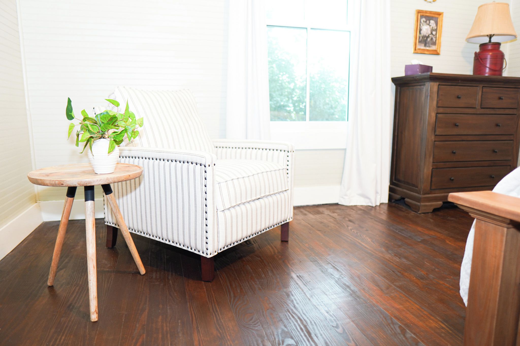 Une pièce comprenant un canapé blanc recouvert de tissu, une table d’appoint en bois avec un pot de plante, une commode en bois et une fenêtre avec des rideaux.