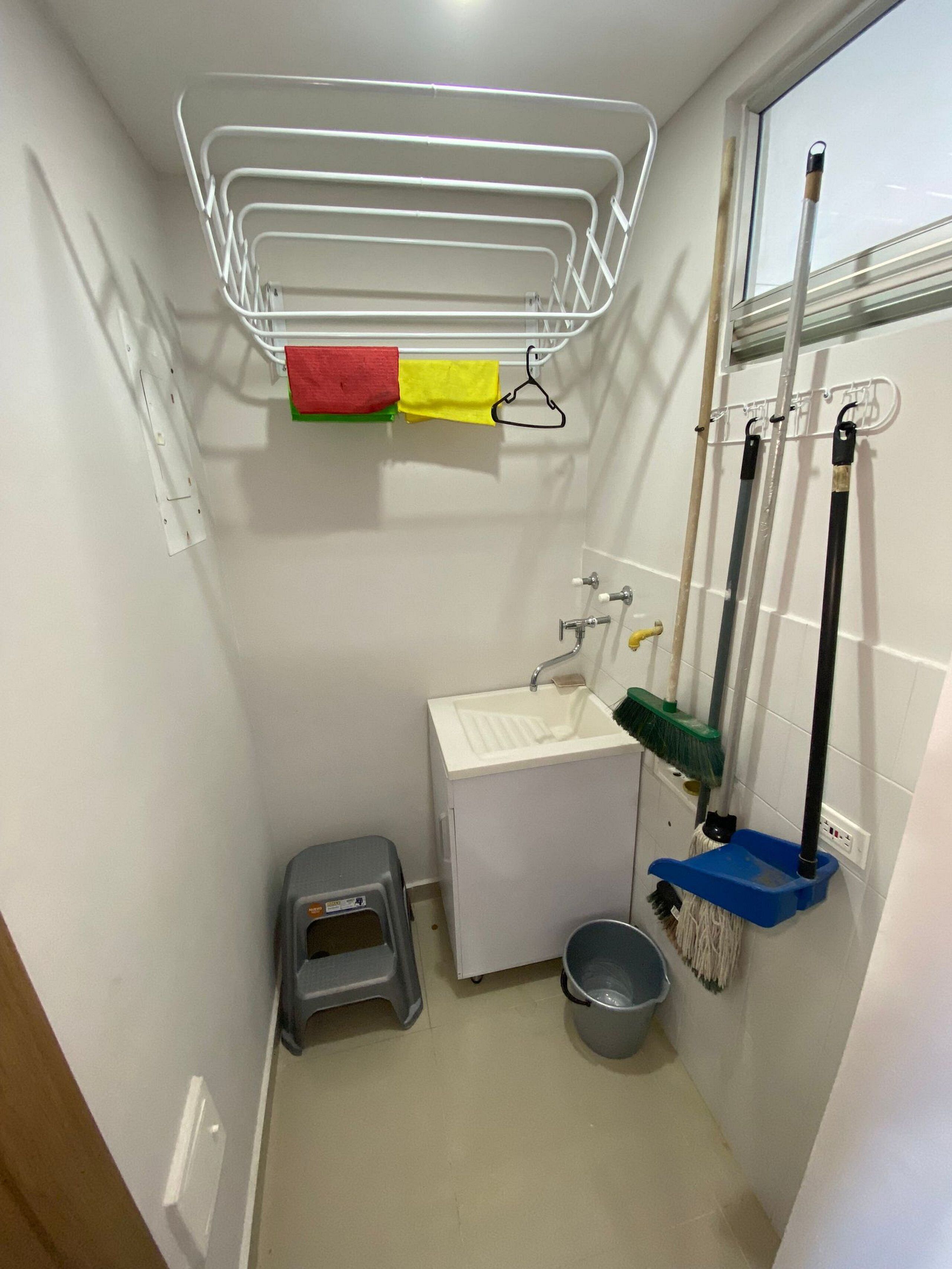 Een kleine badkamer met een wit wastafelmeubel, een spiegel, een handdoekhouder met rode en gele handdoeken, een bezem, een dweil en een kleine prullenbak.
