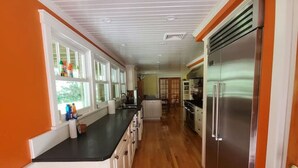 A kitchen with wooden flooring, a bench, a refrigerator, and a window with bottles on the counter. - South Beach | Katama | Outdoor Shower ☀️ 🏖 (Edgartown)