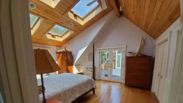 A bedroom with a sloped ceiling, skylights, a bed, a nightstand, a dresser, and a door leading to a balcony. - South Beach | Katama | Outdoor Shower ☀️ 🏖 (Edgartown)