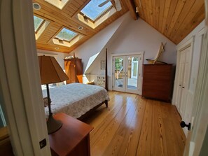 Een slaapkamer met een schuine plafond, dakramen, een bed, een nachtkastje, een dressoir en een deur die leidt naar een balkon.