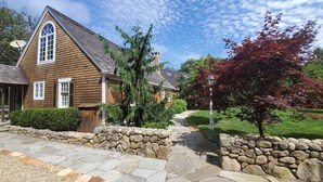 A wooden house with a stone pathway leading to the entrance, surrounded by greenery and a clear sky. - South Beach | Katama | Outdoor Shower ☀️ 🏖 (Edgartown)