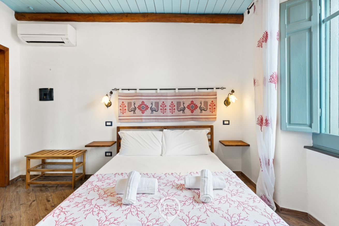 Ein Hotelzimmer mit einem Bett, zwei Nachttischen, einem Holzbeistelltisch, einer Wand-Klimaanlage und einem gemusterten Wandteppich über dem Bett.